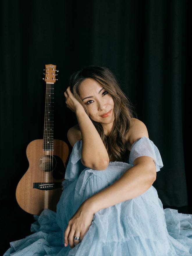 Adlina seated in a blue tulle gown with her acoustic guitar standing behind her
