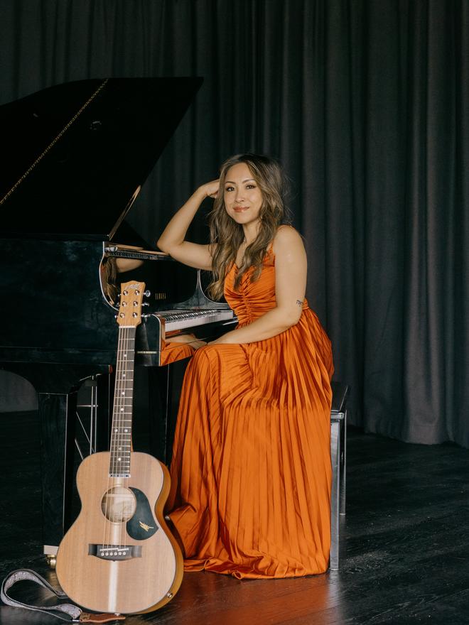 Adlina standing at a grand piano with her acoustic guitar resting against it