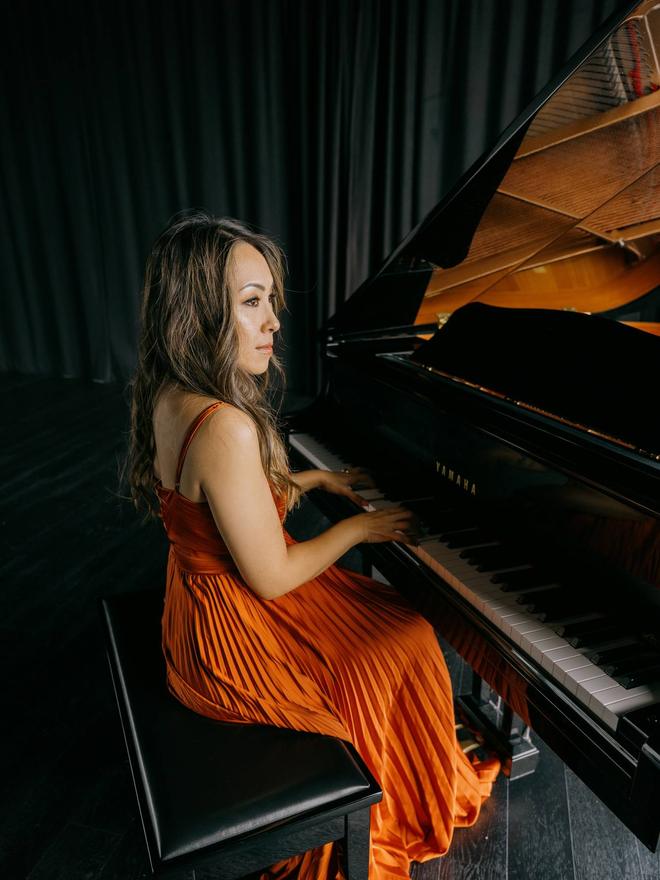 Adlina seated at a grand piano in an amber gown, looking up from the keys