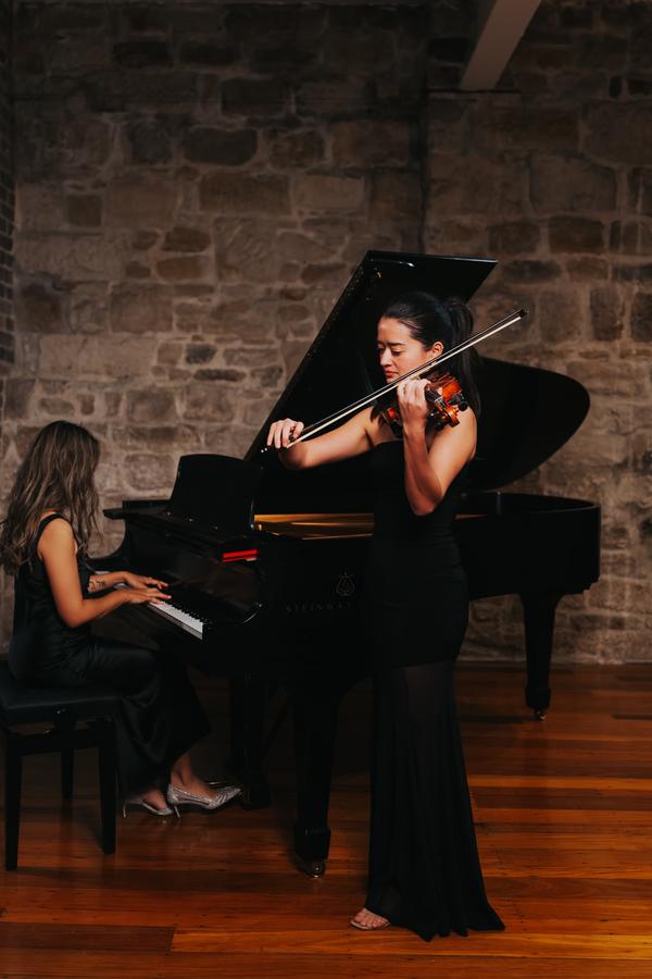 A violinist standing beside a pianist at a grand piano against sandstone