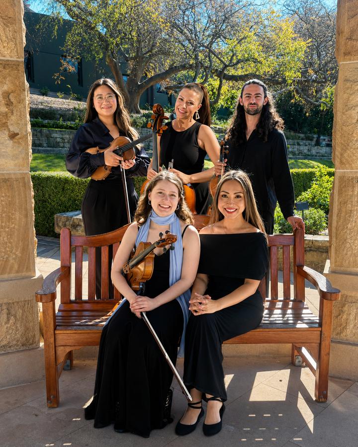 Five musicians in black, seated and standing around a garden bench