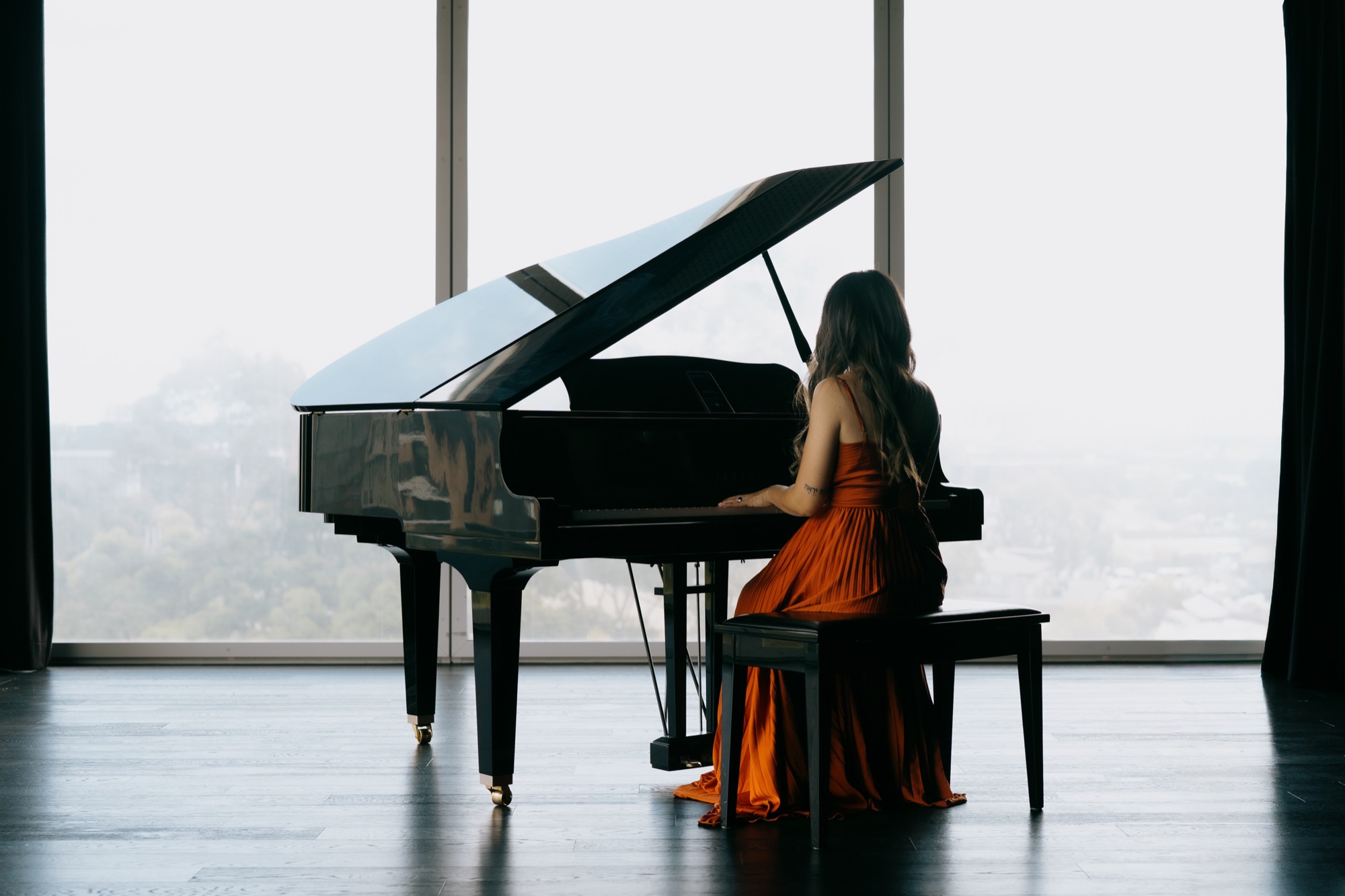 Adlina playing a grand piano in a room overlooking the city through floor-to-ceiling windows
