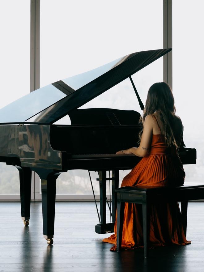 Adlina playing a grand piano beside floor-to-ceiling windows, seen from behind